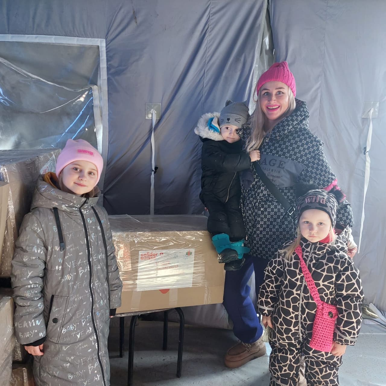 A mother with three children stands near humanitarian aid boxes in a tent camp. The children are dressed in warm winter clothing.
