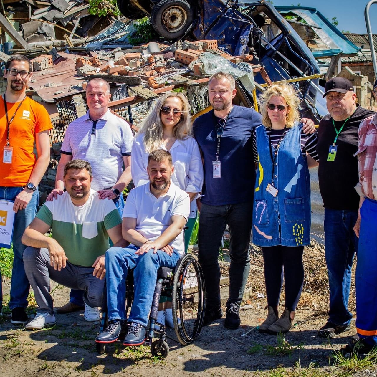A group of people stands behind two men, one of whom is in a wheelchair, against the backdrop of a destroyed building and a blue car damaged during shelling
