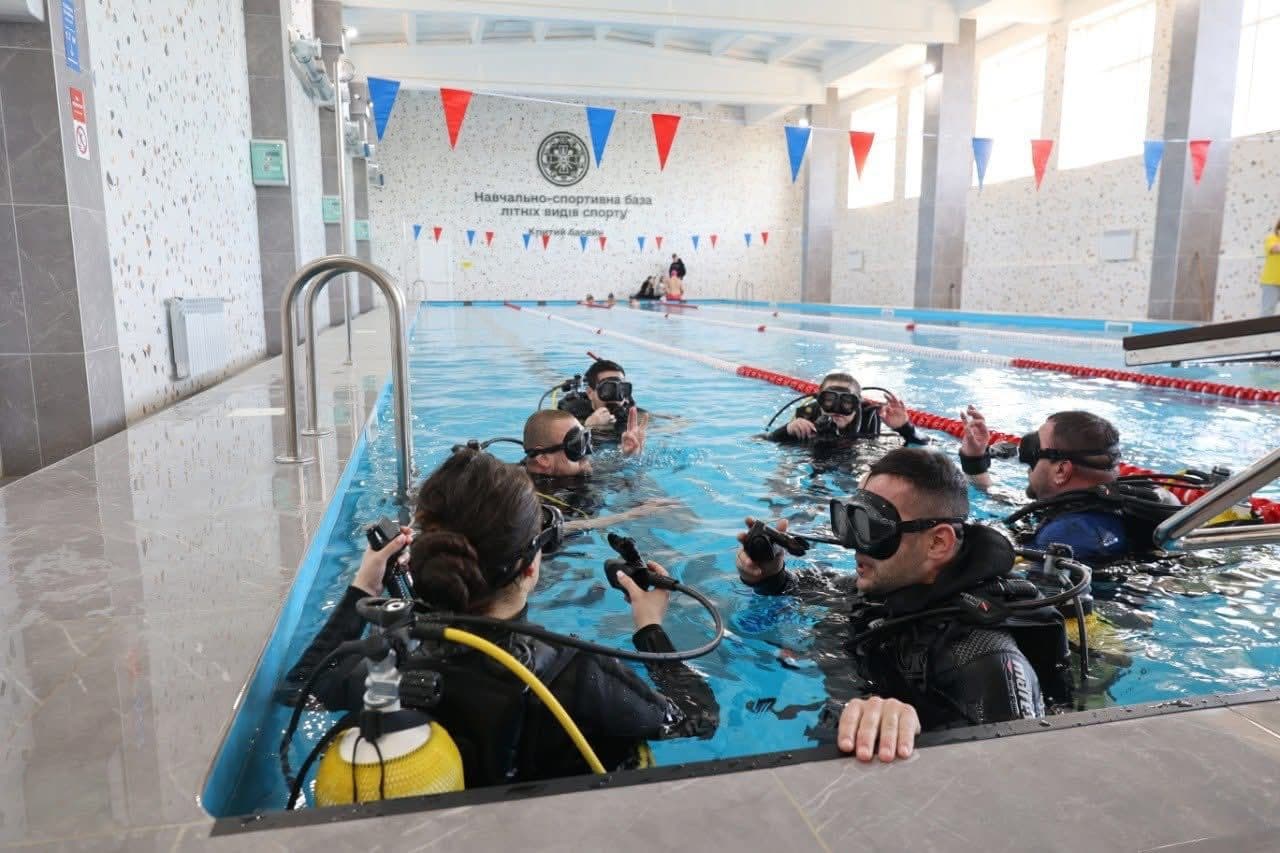 Група людей бере участь у тренуванні з дайвінгу в критому басейні. Учасники вдягнені у повне спорядження: гідрокостюми, маски, трубки та балони з киснем. Вони зібралися біля краю басейну, спілкуються та відпрацьовують сигнали й процедури безпеки. Простір добре освітлений завдяки великим вікнам, стіни прикрашені червоними та синіми трикутними прапорцями. На дальній стіні напис українською: «Навчально-спортивна база. Літніх видів спорту. Плавальний басейн». Сцена демонструє структуроване навчання та підготовку до дайвінгу.