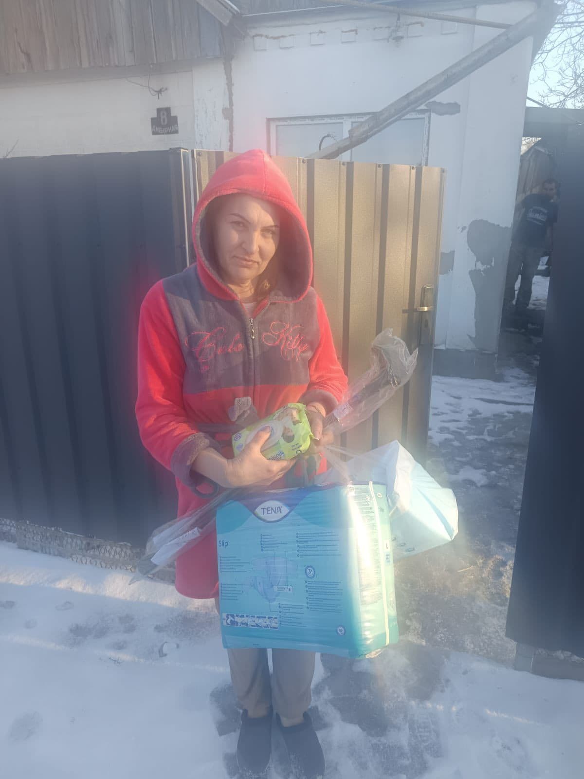 Людина стоїть на вулиці взимку, тримаючи кілька предметів: упаковку підгузків для дорослих TENA Slip, вологі серветки та інші засоби, загорнуті у прозору плівку. Вона одягнена у червоно-сірий худі з написом «Hello Kitty». На задньому плані видно металеву браму та білу будівлю зі слідами зношення й латками на стіні. Інша людина частково видна біля будівлі. Сцена демонструє отримання чи розподіл допомоги у холодному, ймовірно сільському середовищі.