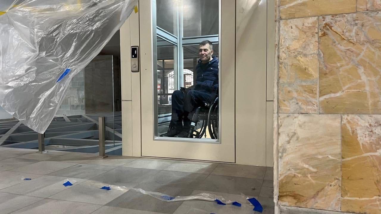 A man in a wheelchair is inside a glass elevator in a modern building. The walls and floor are tiled. A transparent plastic sheet with blue painter’s tape hangs on the left side, indicating ongoing construction or installation. The elevator is designed for accessibility, supporting mobility for individuals with disabilities. The scene highlights inclusive infrastructure in public spaces.