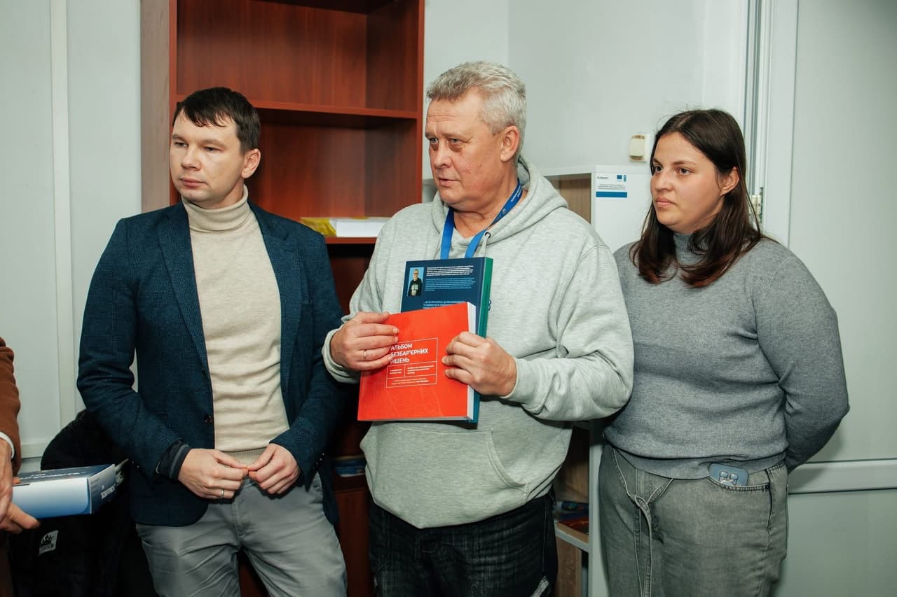 Three individuals stand in an indoor space resembling an office or educational setting. The person in the center holds several books, including a prominent red one titled “Album of Laboratory Solutions.” The others appear to be listening or discussing. Shelves and cabinets are visible in the background. The scene captures a moment of educational or scientific presentation.