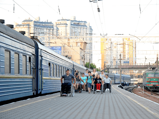 Людина у візку піднімається на потяг за допомогою металевого платформеного підйомника. Потяг стоїть на платформі, а особа одягнена в маску та рукавички. Підйомник має рельєфну металеву поверхню та призначений для допомоги людям із порушеннями мобільності при посадці на потяг. Це зображення підкреслює важливість доступності громадського транспорту для людей з інвалідністю.