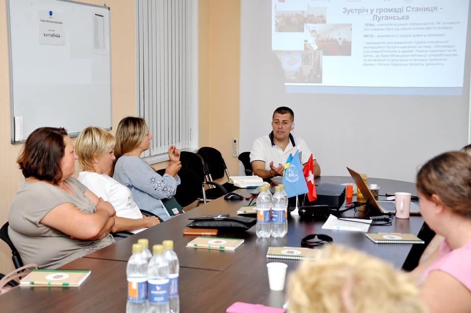A man delivers a presentation to a group of participants. A slide with the meeting topic is displayed on the projector screen, and the Ukrainian and Swiss flags are placed on the table.