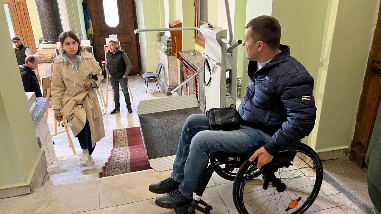 A man in a wheelchair using a stair lift to ascend a flight of stairs in a building. The stair lift is a mechanical device designed to assist individuals with mobility impairments. The man wears a blue jacket and jeans and has a black bag on his lap. Several people are in the background, including one person using crutches and others standing near the entrance. The building has a marble floor, a red carpet on the stairs, and wooden doors, emphasizing efforts to accommodate individuals with disabilities.