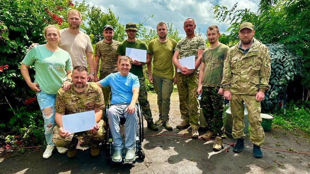 A group of eleven people poses outdoors amid greenery under a cloudy sky. Most wear military or camouflage clothing. One person in a light shirt and pants sits in a wheelchair. Three individuals hold sheets of paper, likely certificates. Equipment and containers are visible in the background. The scene conveys recognition, solidarity, and camaraderie, possibly linked to military service or a training event.