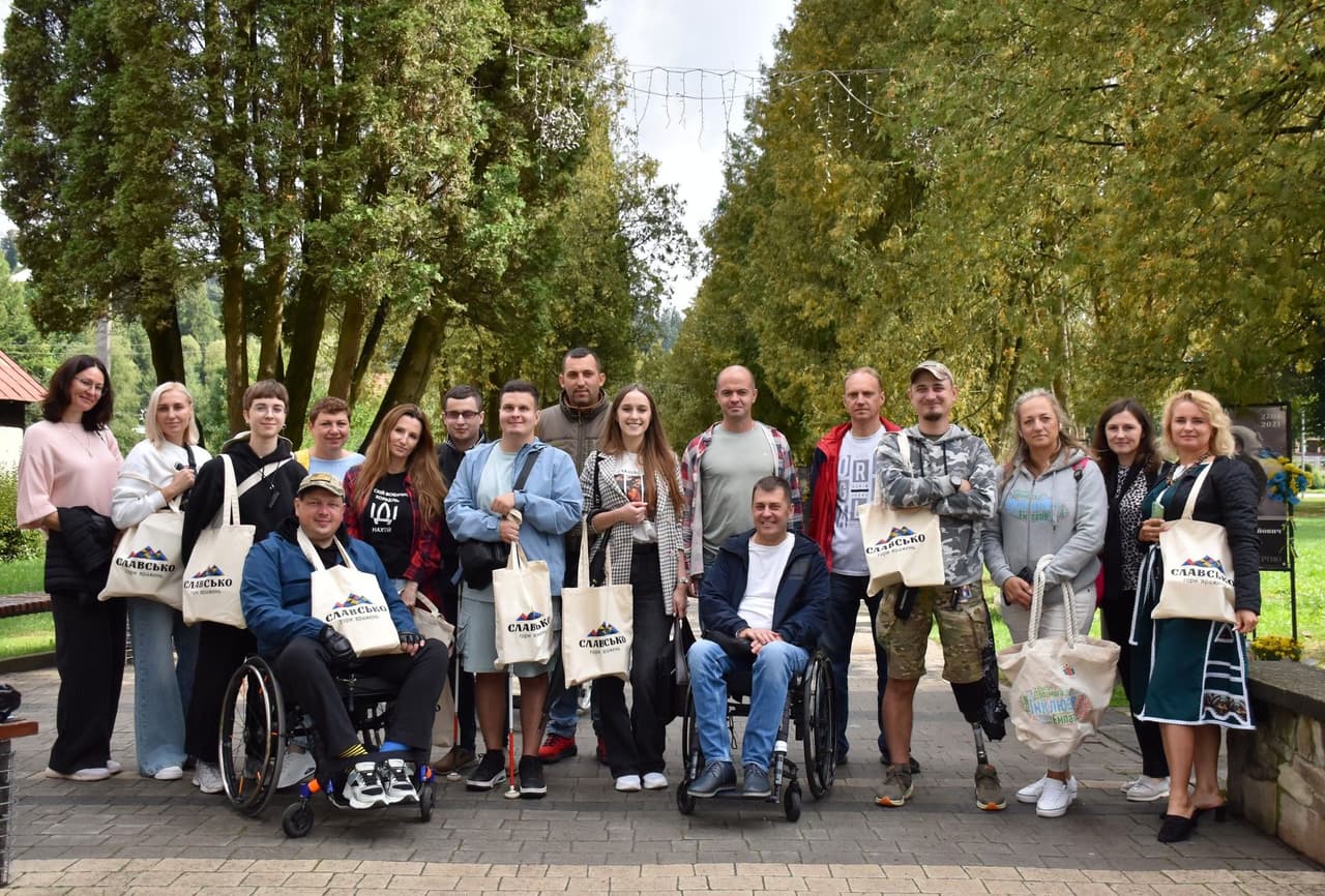 Група з 18 людей позує на відкритому просторі, стоячи на вимощеній доріжці серед високих зелених дерев. Серед учасників — двоє осіб у візках, що підкреслює інклюзивність і доступність. Багато хто тримає екоторби з написом “СЛАВА” та логотипом, що свідчить про участь у спільному заході або кампанії. У групі — чоловіки й жінки в повсякденному одязі. Атмосфера передає згуртованість, видимість і спільну підтримку.