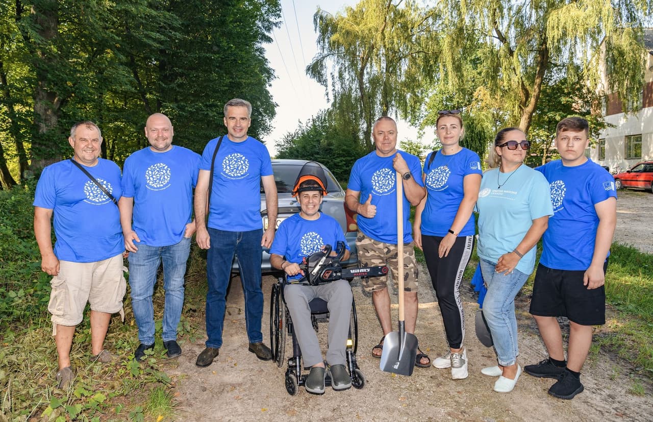 The image shows a group of nine people standing outdoors, possibly in a park or wooded area. They are all wearing blue t-shirts with a white circular logo on the front. One person is seated in a wheelchair holding a piece of equipment, and another person is holding a shovel. The group appears to be participating in a community or volunteer event, possibly related to outdoor work or environmental conservation.