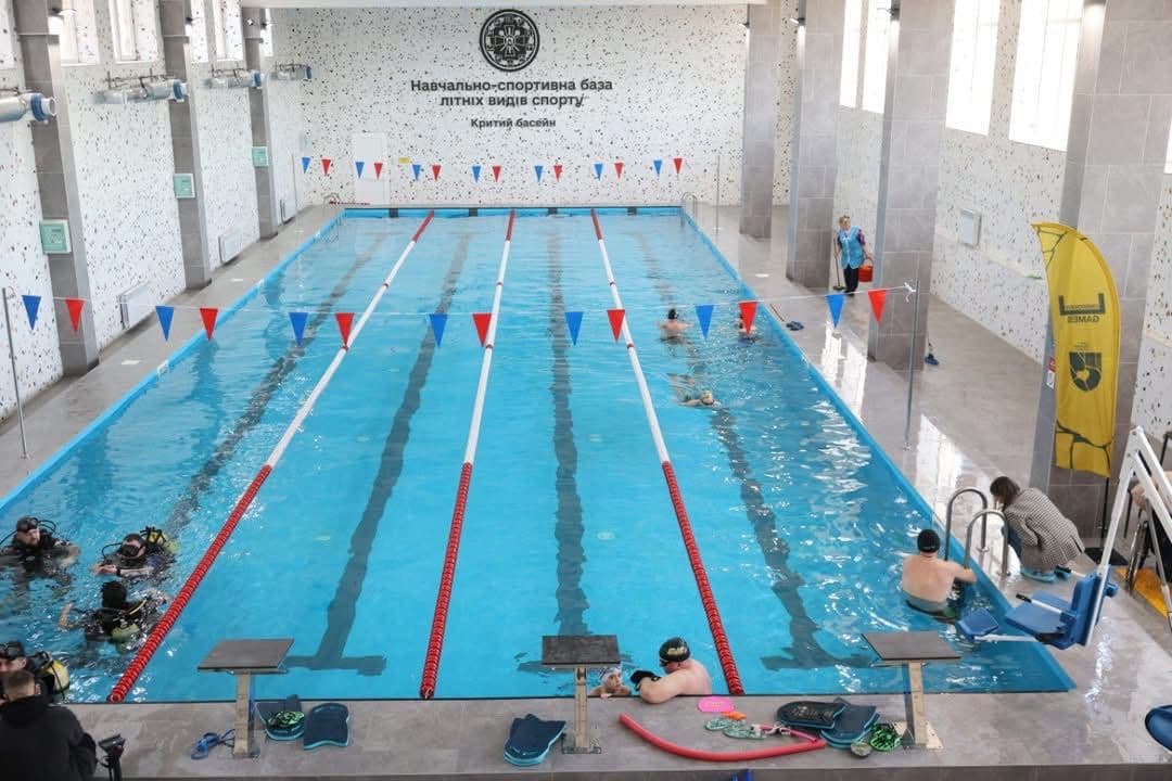 Критий басейн із кількома доріжками, розділеними буйками. У воді перебувають кілька людей, частина з них у спорядженні для дайвінгу, що свідчить про навчальне чи тренувальне заняття. Простір добре освітлений завдяки великим вікнам. На дальній стіні видно напис українською: «Навчально-спортивна база літніх видів спорту. Критий басейн» та круглу емблему. Над доріжками натягнуті червоні й сині трикутні прапорці. Праворуч — жовтий вертикальний банер із написом «GAMES» та зеленим щитом. На бортику розташоване обладнання для плавання й дайвінгу: дошки, ласти, нудли. Сцена демонструє багатофункціональне використання басейну для плавання та підводного навчання.