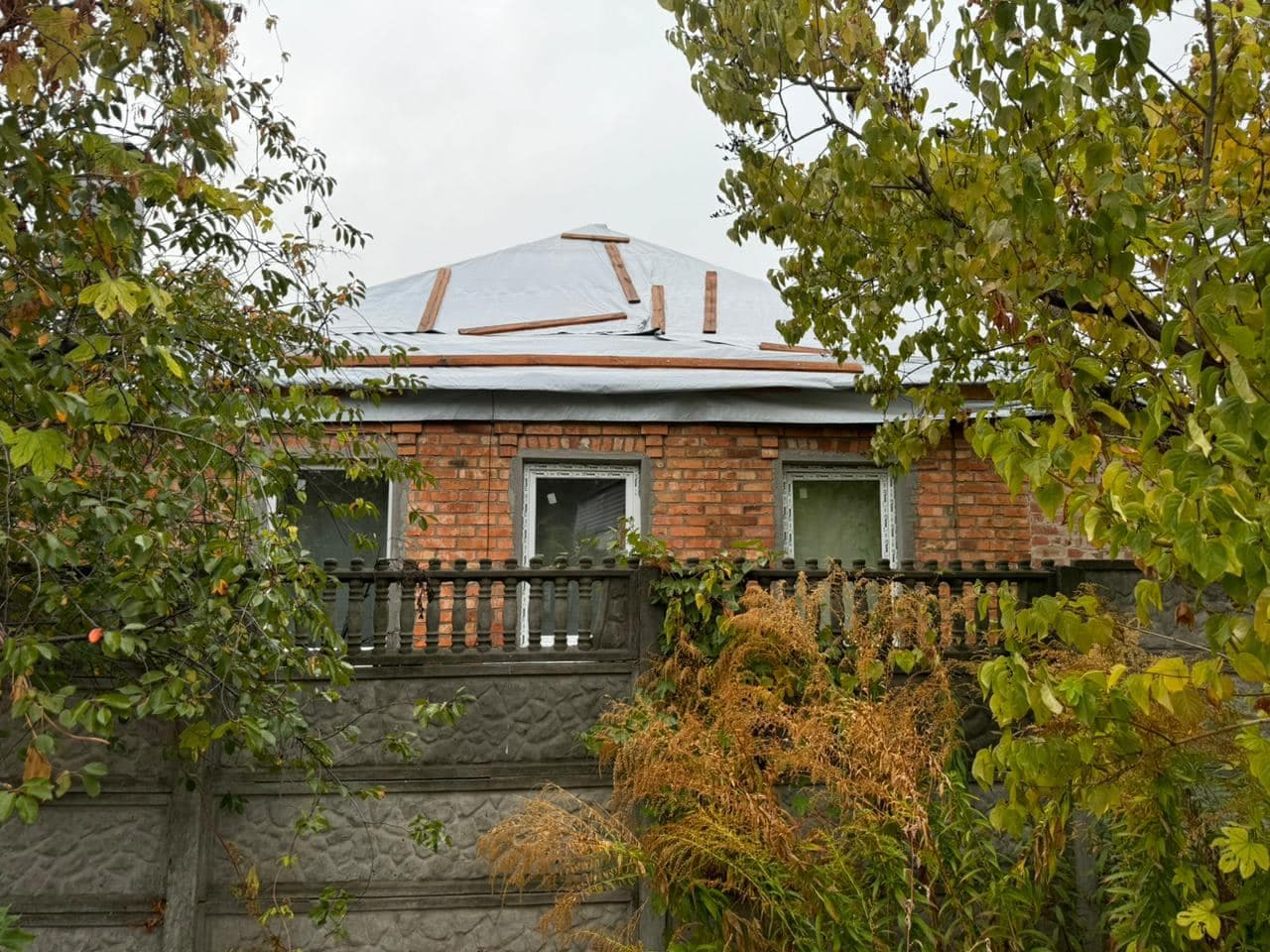A brick house behind a concrete fence with trees growing in front of it. The roof, damaged by shelling, is covered with gray construction fabric, with wooden planks placed on top.