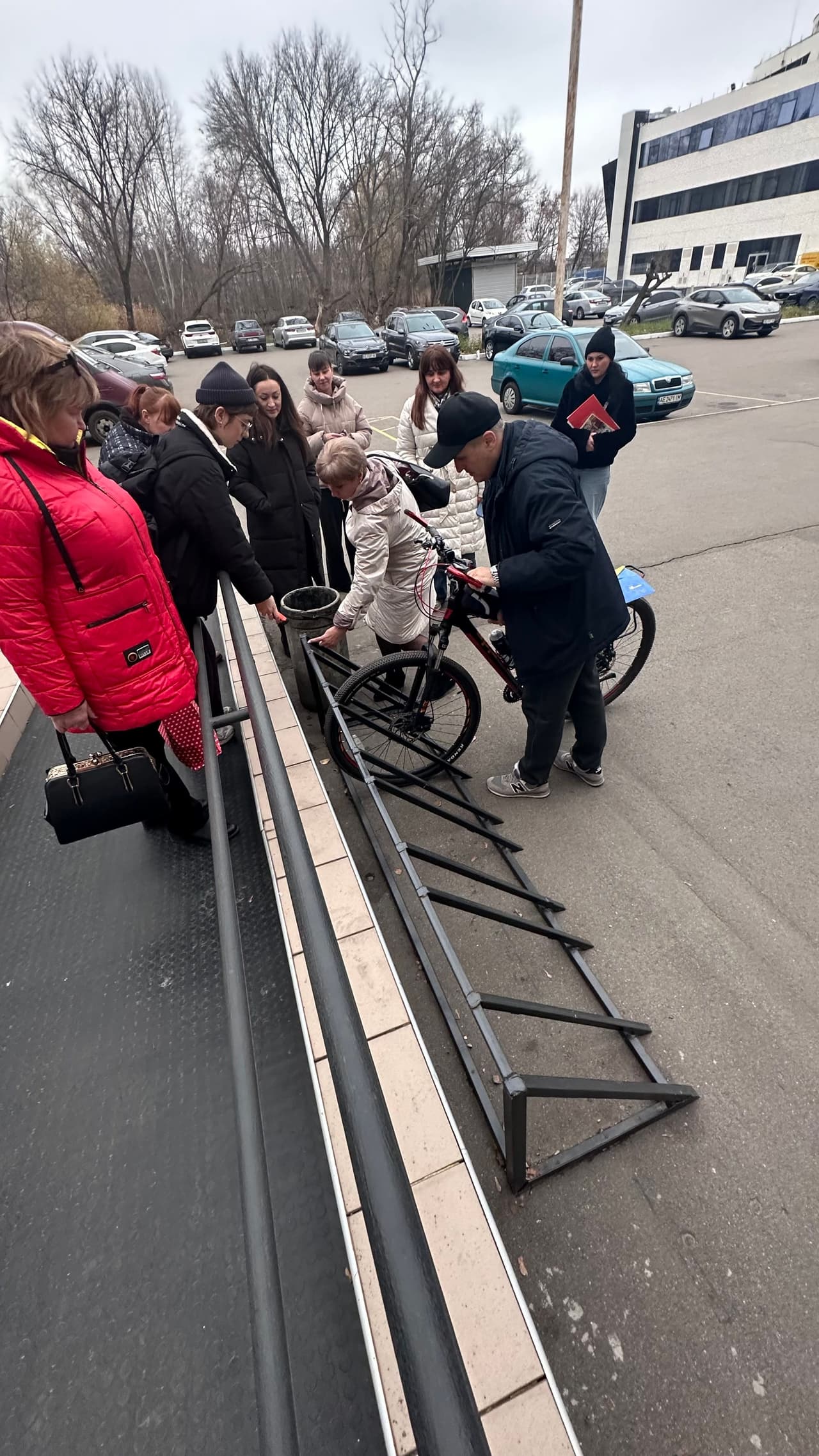 «Група учасників тренінгу проводить практичний огляд зовнішнього простору біля входу до будівлі. На передньому плані — пандус із гумовим протиковзким покриттям та металевими поручнями. Поруч розташована металева рампа для велосипедів, встановлена на рівні проїзної частини. Учасники аналізують відповідність елементів доступності вимогам ДБН, звертаючи увагу на розміщення пандуса, ухил, наявність контрастних позначень і потенційні перешкоди на маршруті.»