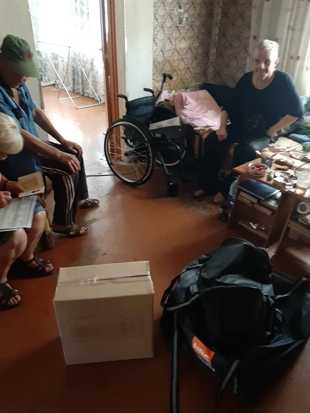 An elderly person sits on a sofa in a room where other people are also present. A wheelchair is nearby, and on the floor, there is a box of humanitarian aid and a black bag with the NRC logo. One of the individuals is holding documents, possibly for aid registration.