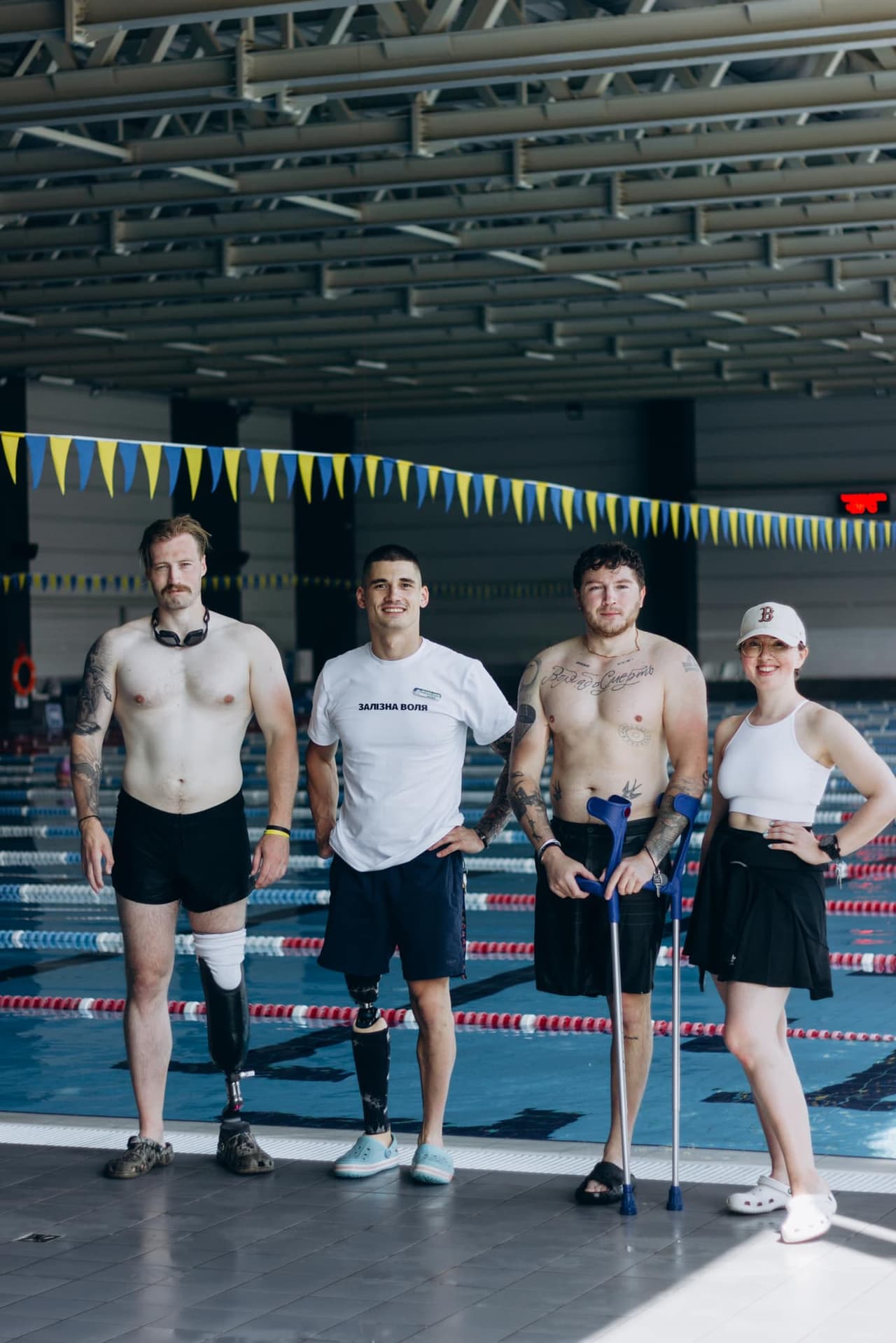 Three men with leg prosthetics and crutches, along with a girl, against the backdrop of a swimming pool.