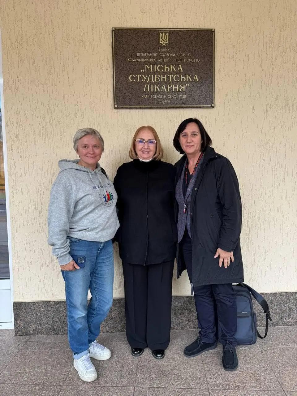 Three women standing together in front of a beige wall with a plaque that reads 'Міська студентська лікарня' (City Student Hospital) in Ukrainian. The woman on the left is wearing a gray hoodie and jeans, the woman in the center is in black clothing, and the woman on the right is in a black jacket with a backpack next to her.