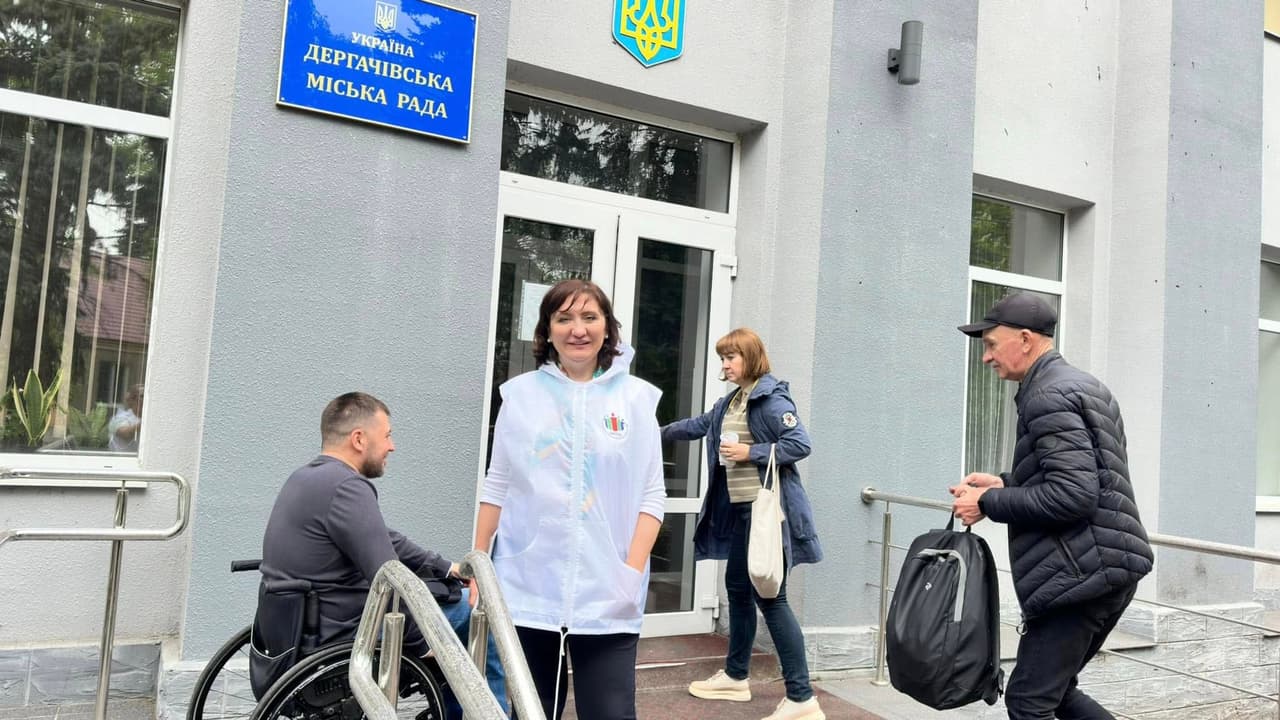 На зображенні зображено вхід до будівлі з написом українською мовою "УКРАЇНА ДЕРГАЧІВСЬКА МІСЬКА РАДА," що означає "Україна Дергачівська міська рада." На передньому плані є четверо людей: одна людина у інвалідному візку, одна людина у білій жилетці стоїть перед входом, ще одна тримає чашку та відкриває двері, а інша носить рюкзак і рухається до входу. Будівля має сірий фасад із великими вікнами та пандусом для доступу на візках. Зображення підкреслює доступність та вхід до урядової будівлі.