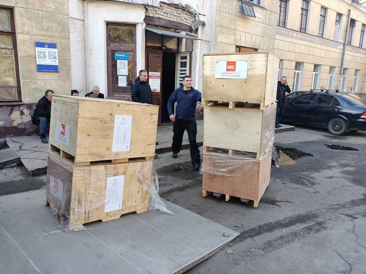 Delivery of humanitarian aid from the Norwegian Refugee Council (NRC). Two men are moving large wooden crates wrapped in plastic, against the backdrop of a run-down building with peeling walls. A few people are sitting nearby, while another man stands by a black car, talking on the phone. The scene highlights the difficulties faced by the community and the importance of receiving aid.