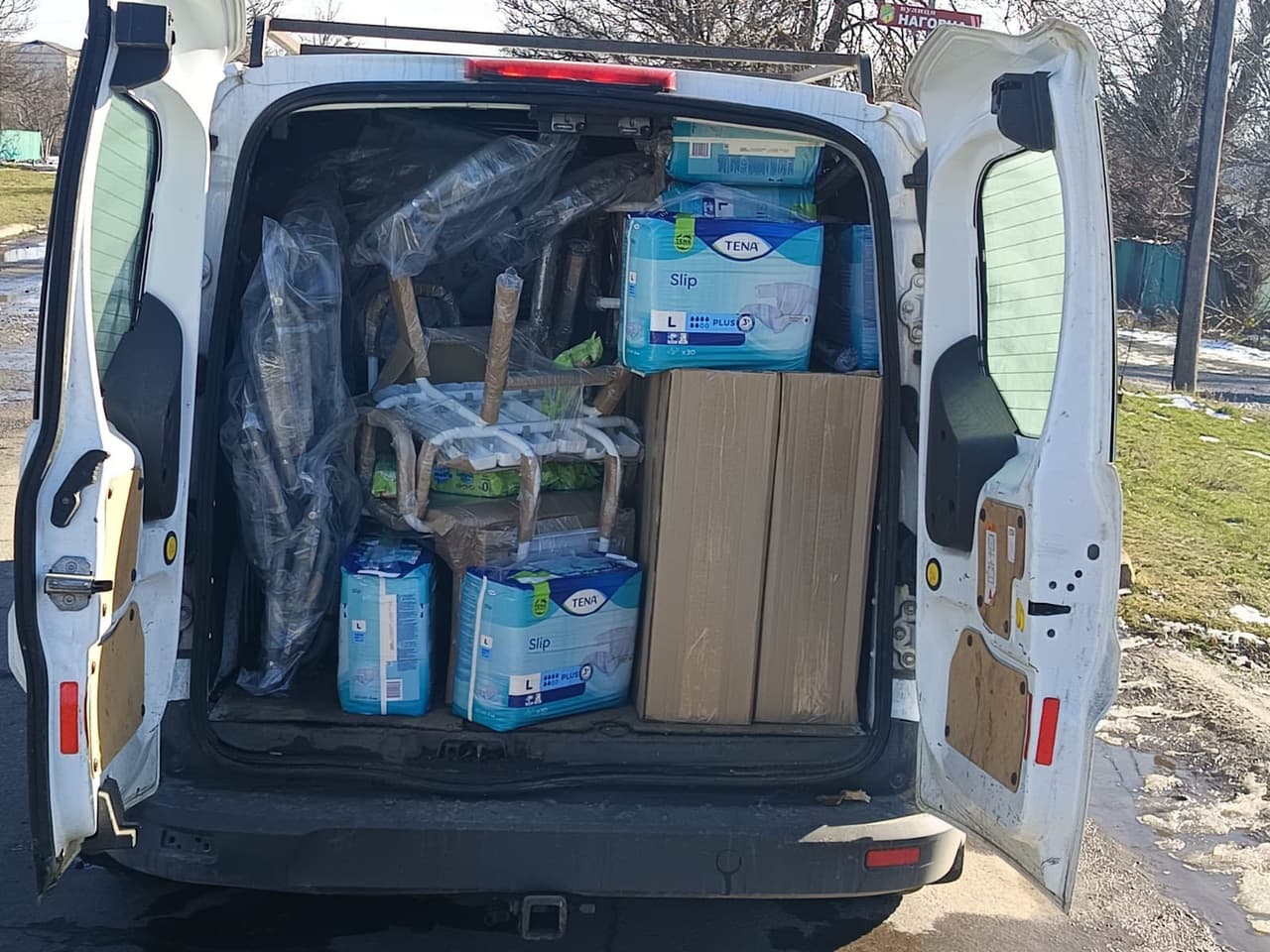 Відкрита задня частина білого фургона, заповненого різними речами. На передньому плані видно кілька упаковок підгузків для дорослих TENA Slip із позначками «L» та «Plus», складених одна на одну. Усередині також великі картонні коробки та загорнуті у захисну плівку предмети, схожі на меблі чи медичне обладнання. Автомобіль припаркований на вулиці, де видно сніг і траву, що свідчить про холодну пору року. Сцена демонструє доставку чи транспортування медичних засобів для догляду.