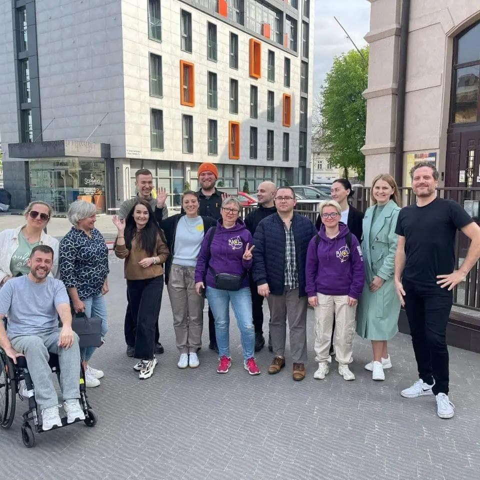Group photo of a diverse team of men and women, including individuals with disabilities, standing outdoors in front of a modern building. Some participants are wearing casual clothes, while others are dressed in purple sweatshirts with a logo. Everyone is smiling, showcasing a positive and inclusive atmosphere.