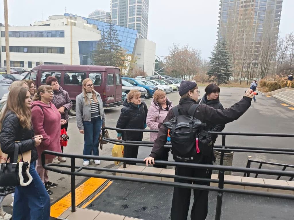A group of people gathered outdoors near a modern building. One person with a backpack holds an object and addresses the group, possibly leading a presentation or tour. Attendees wear casual cool-weather clothing. In the background are a van, parked cars, trees, and tall buildings, suggesting an urban educational or community event.