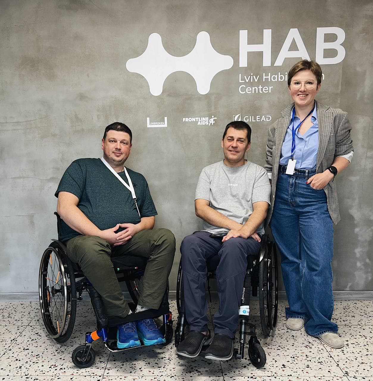Two men are sitting in wheelchairs, with a woman standing next to them, against the backdrop of the “HAB” logo.