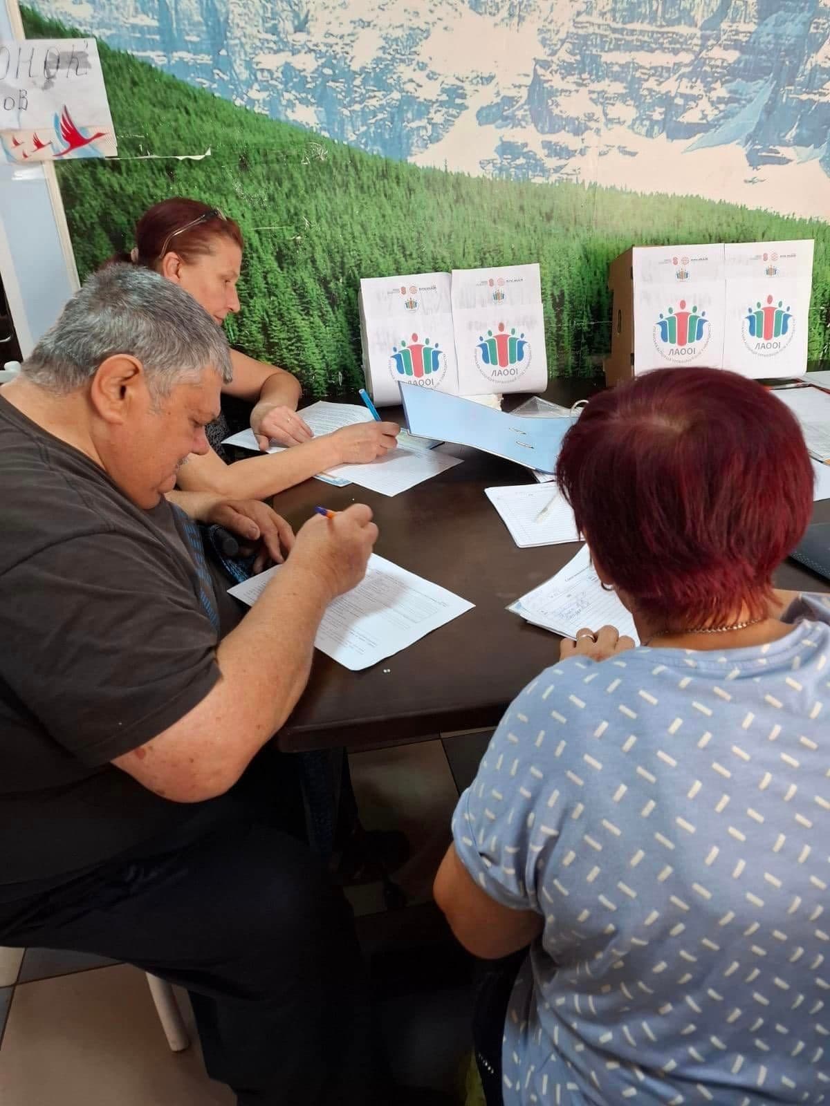 Two women and a man are sitting at a table, writing on paper.