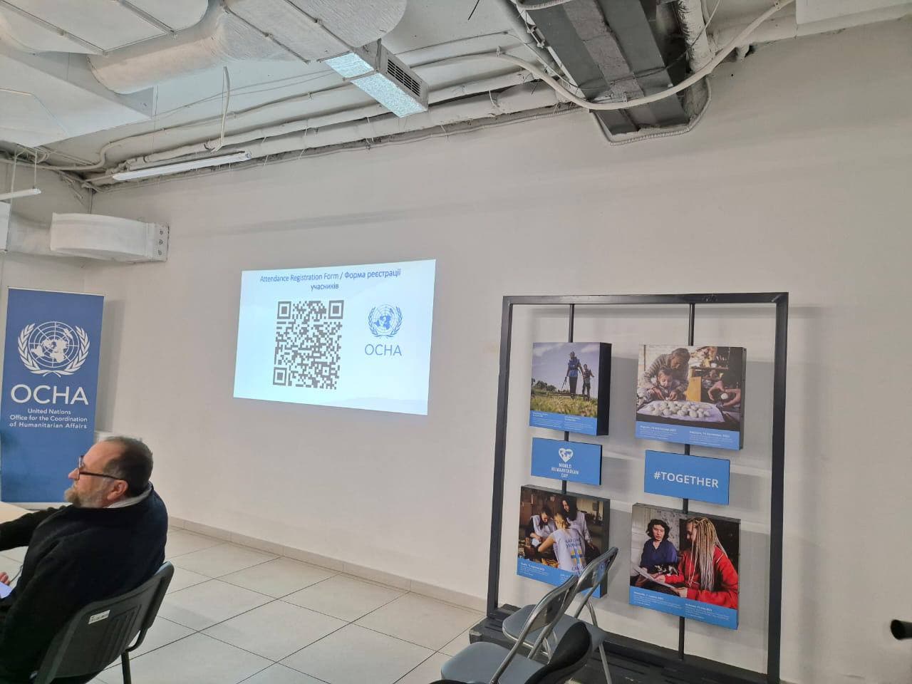 Конференція в приміщенні з елементами, пов'язаними з Офісом ООН із координації гуманітарних питань (OCHA). Ліворуч розташований банер із логотипом OCHA та текстом, що ідентифікує організацію. На стіні проектується зображення з QR-кодом, логотипом OCHA та текстом \"Форма реєстрації учасників.\" Праворуч стоїть стенд із зображеннями та описами гуманітарних заходів, зокрема текст \"#RAZOM.\"