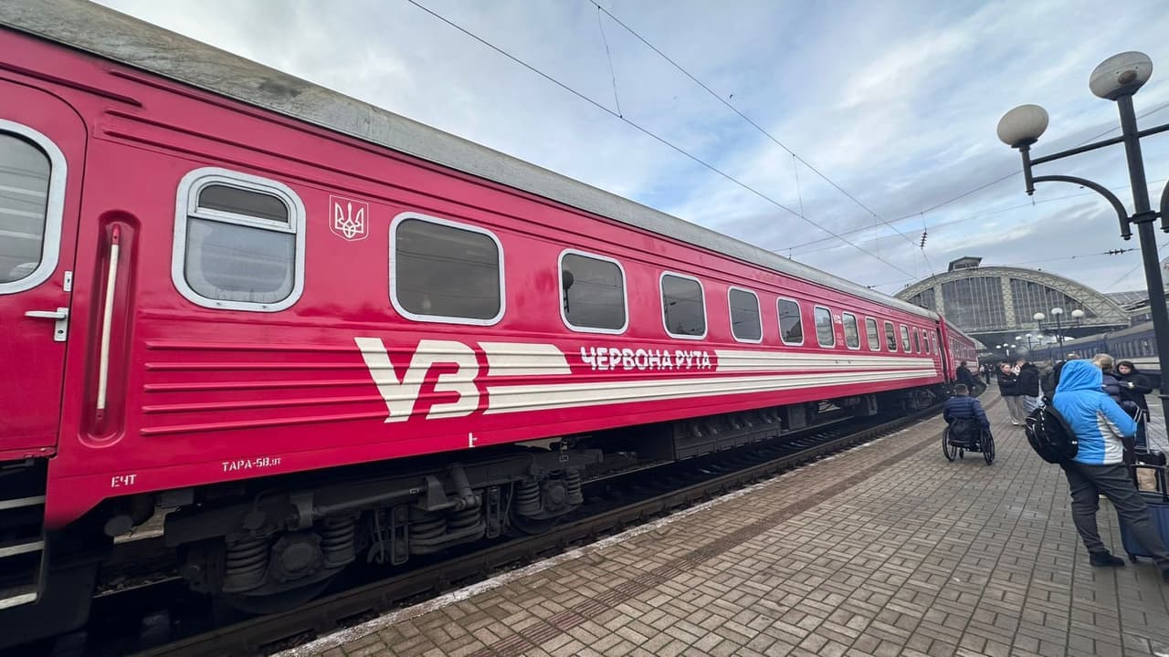 Яскраво-червоний пасажирський поїзд із написами «УЗ» і «Червона рута» стоїть на платформі вокзалу. Біля вагона — люди з багажем, зокрема особа на інвалідному візку. На задньому плані — аркова будівля вокзалу, електричні дроти та світильники. Сцена демонструє доступність залізничного транспорту в Україні.