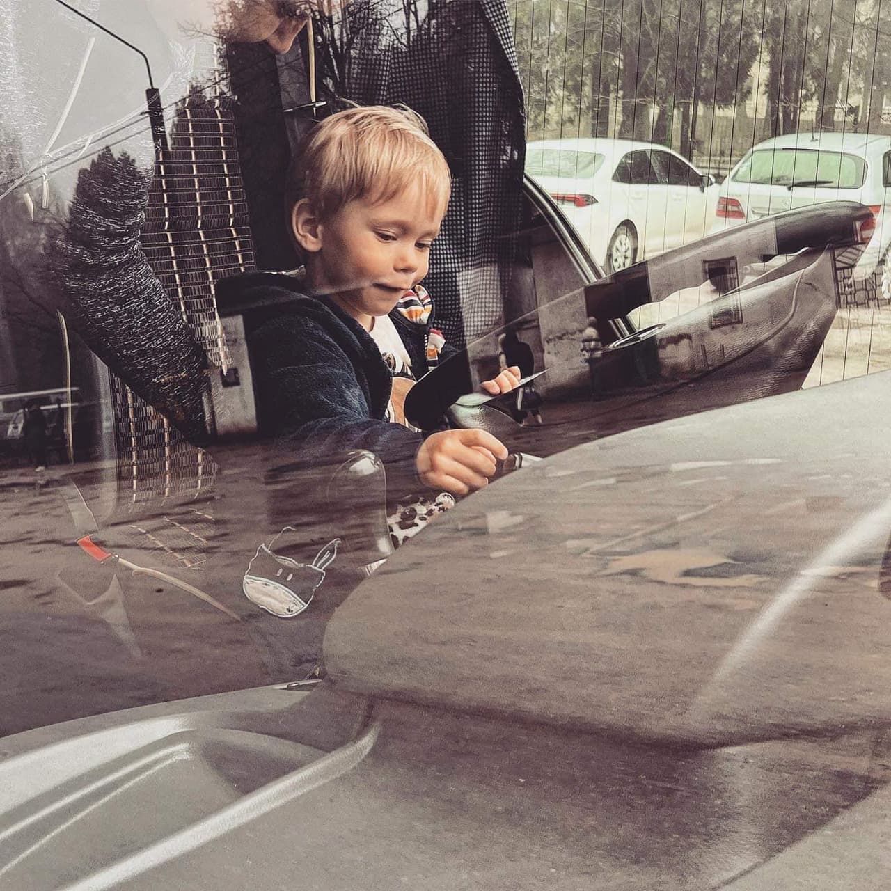 На зображенні видно малу дитину, що сидить на водійському сидінні автомобіля і тримається за кермо. Дитина дивиться вперед, а зображення зроблене ззовні автомобіля через лобове скло. На лобовому склі видно відображення навколишнього середовища, включаючи припарковані автомобілі та дерева. Обличчя дитини розмиті для забезпечення приватності. Це зображення цікаве тим, що захоплює момент, коли дитина імітує водіння, що є поширеною поведінкою у грі.