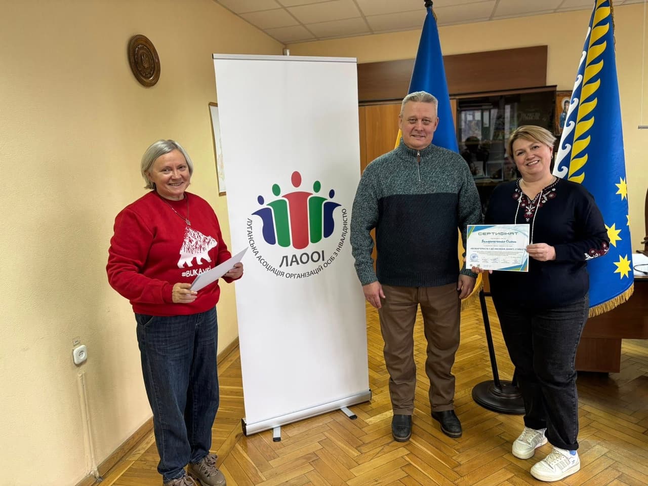 Three people pose indoors on a wooden floor. On the left is a person in a red sweatshirt with a white bear graphic and the hashtag “#BUKAVINA.” In the center stands a person in a dark green sweater. On the right, a woman in floral-embroidered dark clothing and white sneakers holds a certificate labeled “CERTIFICATE Issued to Svitlana.” Behind them are two flags, including the Ukrainian national flag, and a banner from the Luhansk Association of Organizations of Persons with Disabilities (LAOOI) featuring stylized human figures. The scene reflects a moment of recognition or program completion related to inclusion or support.