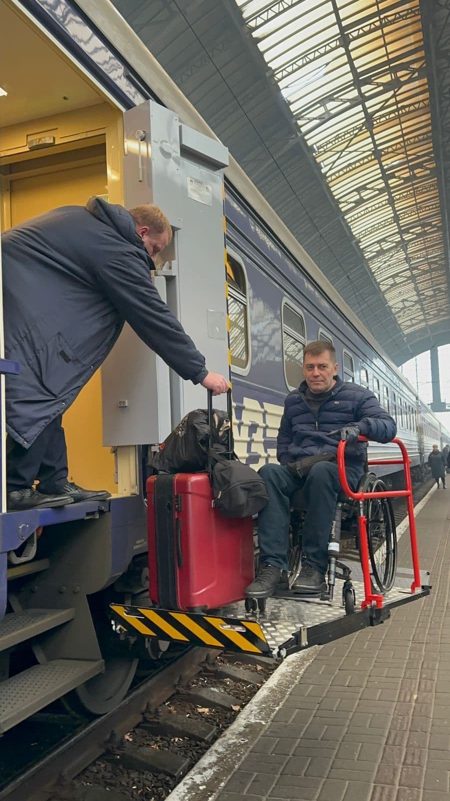 Чоловіка на інвалідному візку підіймають на платформу підйомника, щоб потрапити до вагона поїзда. Працівник допомагає з багажем і керує механізмом. Сцена відбувається на вокзалі під скляним дахом. Зображення демонструє доступність громадського транспорту для людей з порушеннями мобільності.