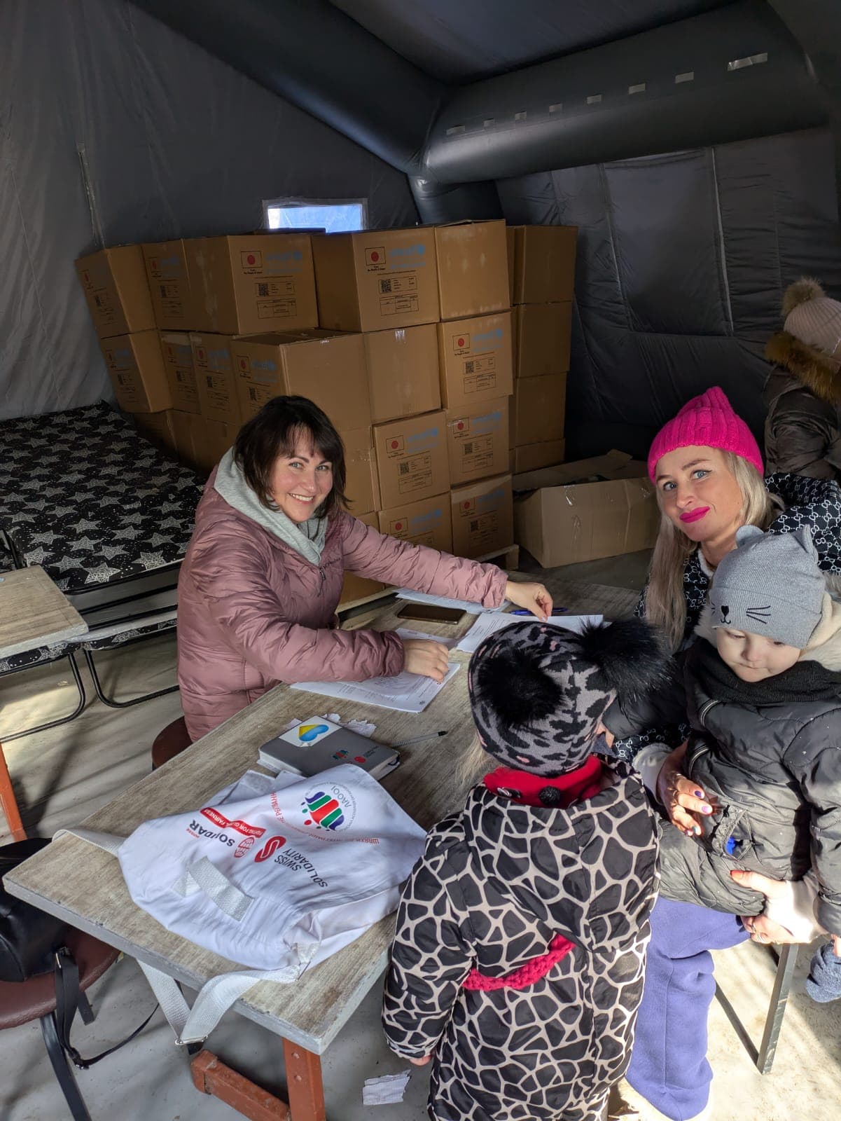A woman in warm clothing sits at a table in a tent, distributing humanitarian aid. In front of her, a mother with children is receiving assistance. Behind them, stacks of aid boxes are visible.