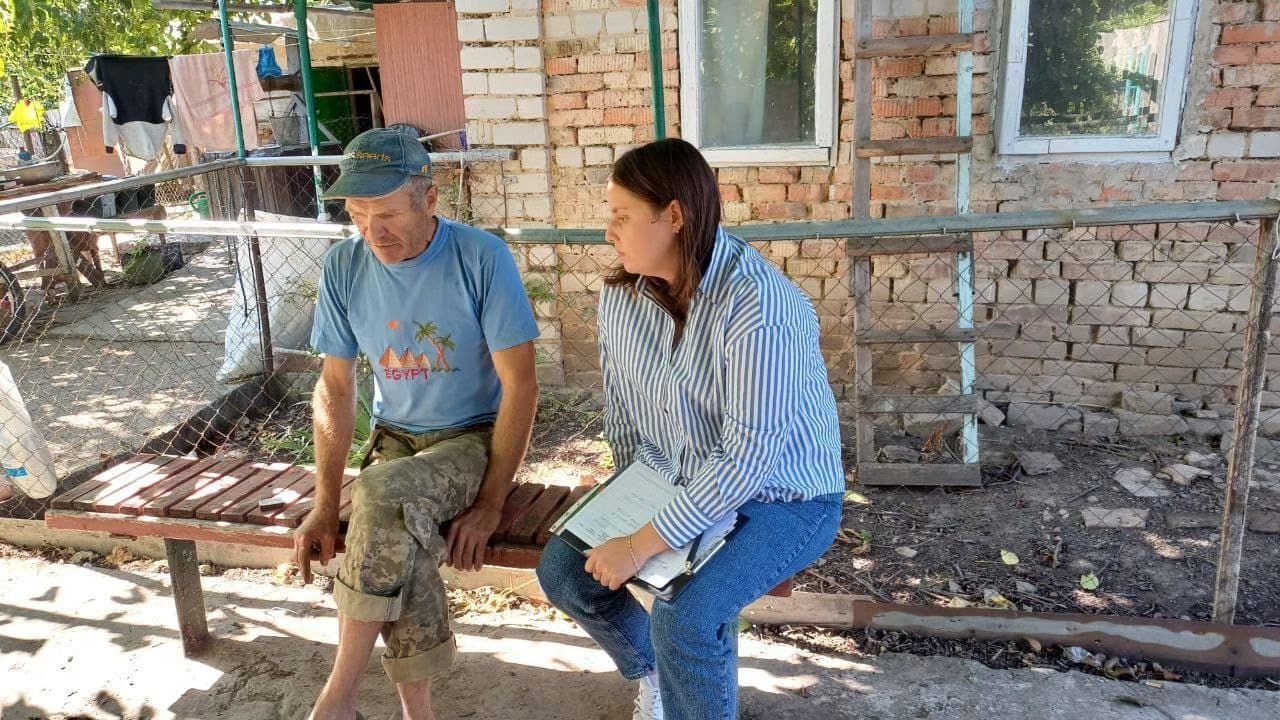 Two individuals sit on a wooden bench outside a brick building. The person on the left, an older adult, wears a blue graphic t-shirt, camouflage pants, and a blue cap. The person on the right, dressed in a blue and white striped shirt and jeans, holds a clipboard and pen, appearing to conduct an interview or conversation. The setting includes a fenced area, dirt ground with scattered leaves, and a backdrop of brick walls and windows, suggesting a quiet rural or residential environment.