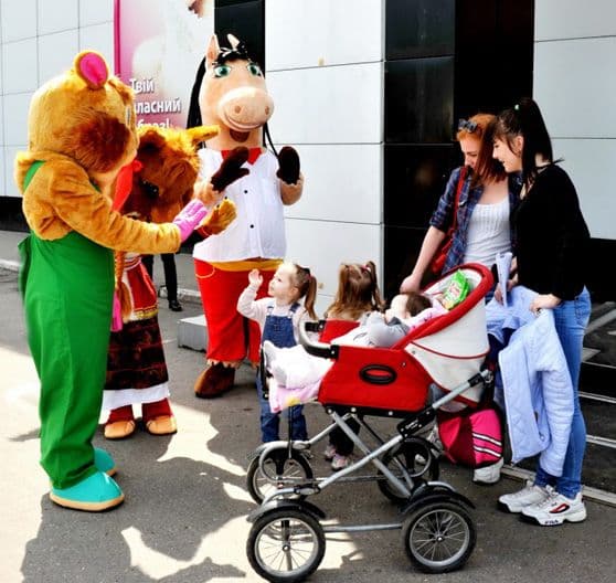 Outside a building, children interact with entertainers dressed as animals – a squirrel, a bear, and a horse. Two little girls excitedly reach out to the characters, while their mothers with a stroller watch with smiles. The atmosphere is joyful and friendly.