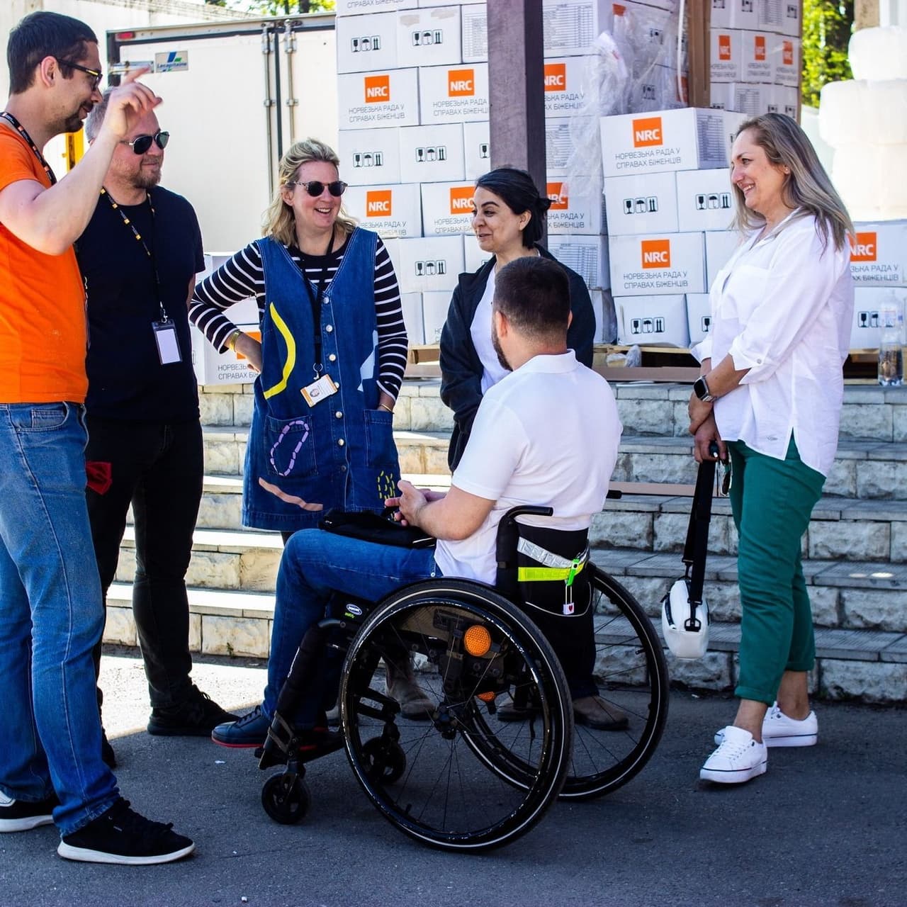 Three women and two men are standing next to a man in a wheelchair, with cardboard boxes featuring an orange logo in the background.