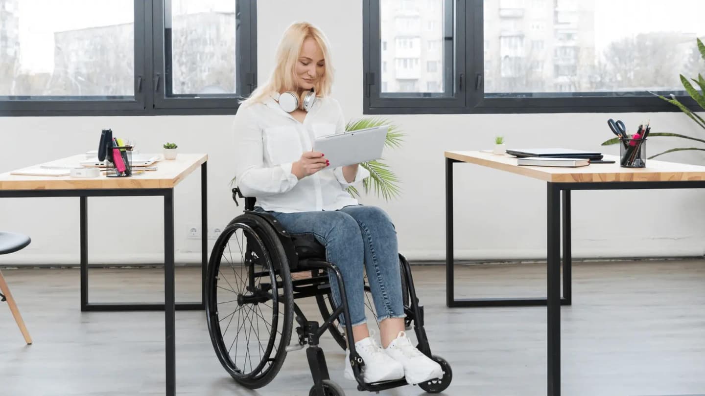 Людина в інвалідному візку сидить у сучасному офісі. Вона одягнена у білу сорочку, джинси та білі кросівки, на шиї навушники. У руках тримає планшет і дивиться на нього. Офіс добре освітлений, із великими вікнами та видом на будівлі зовні. Видно два дерев’яні столи з чорними металевими ніжками, на яких розташовані канцелярські матеріали, блокноти та маленькі вазони. Сцена демонструє інклюзивне та професійне робоче середовище.