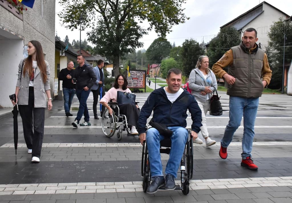 A group of people crosses a street at a pedestrian crosswalk in an urban setting. Among them are two individuals using wheelchairs. The participants are casually dressed, some engaged in conversation. Buildings, trees, and a signboard are visible in the background. The scene highlights inclusivity and the accessibility of public infrastructure for people with mobility impairments.