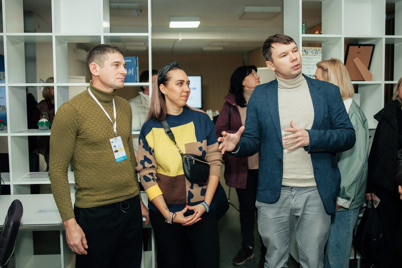 A group of people engages in conversation in a modern office space with white shelving units holding books, documents, and decor. One man gestures as if explaining something. Participants wear business or casual attire, and one person has a name badge. The scene reflects a professional exchange, possibly during a tour, workshop, or networking event.