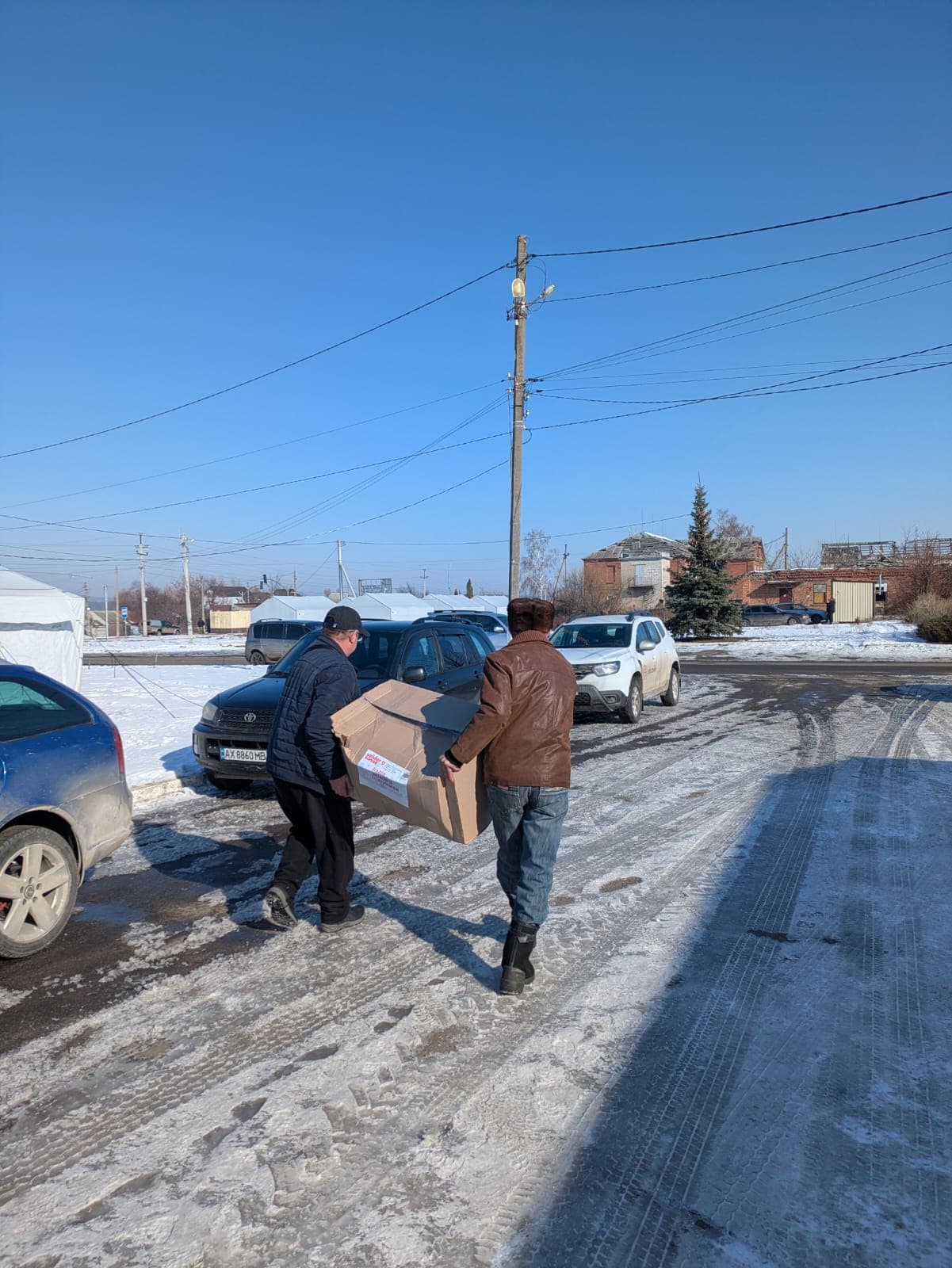 Two men carry a box of humanitarian aid along a snowy road. Parked cars and a tent camp are visible in the background.