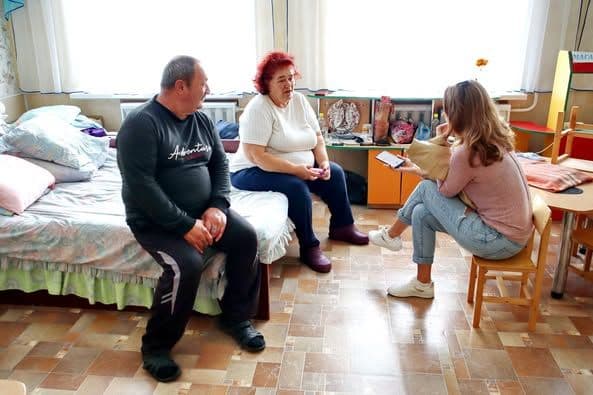 The image shows three people in a room. Two of the people are sitting on a bed, while the third person is sitting on a small chair facing them. The person on the left is wearing a black long-sleeve shirt and black pants, while the person in the middle is wearing a white shirt and dark pants. The person on the right is wearing a pink sweater and blue jeans, holding a notepad and pen, appearing to be taking notes or conducting an interview. The room has a bed with pillows and a blanket, a small table, and various items on a shelf against the wall. The floor is covered with a patterned linoleum.