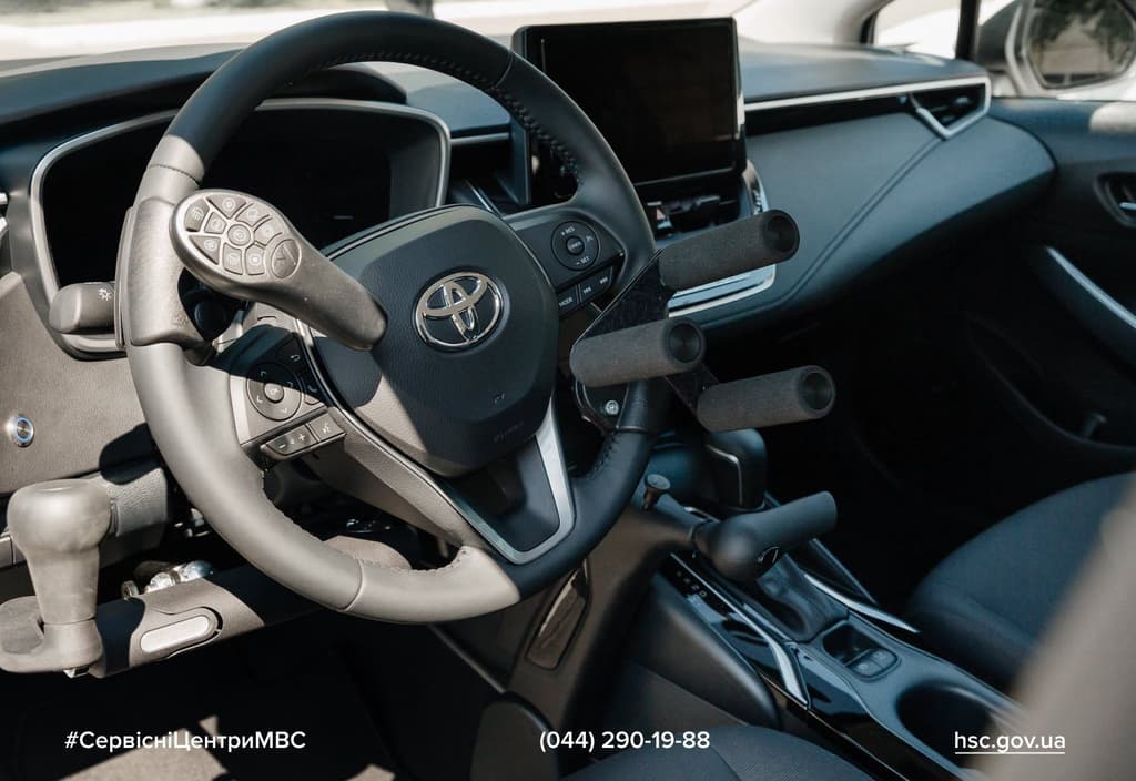 Інтер’єр модифікованого автомобіля Toyota з адаптованими елементами керування для водіїв з порушеннями рухливості. На кермі — спеціальні важелі та ручки з м’якими накладками. Видно сенсорний дисплей на панелі приладів і важіль перемикання передач. Зображення демонструє приклад доступного транспорту.