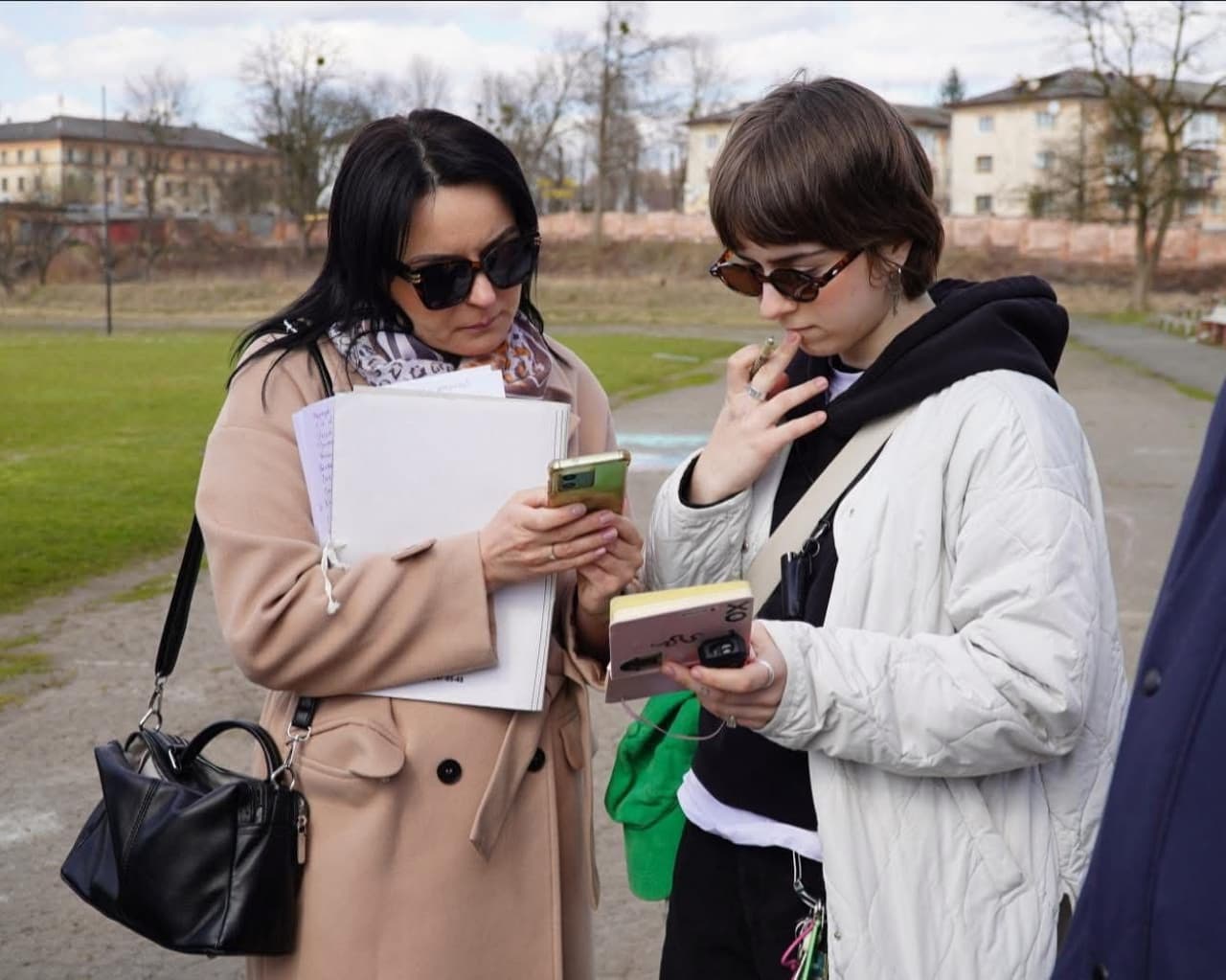 На зображенні двоє людей стоять на вулиці та дивляться у свої телефони. Одна людина в бежевому пальті тримає стос паперів або зошитів із рукописними нотатками. Інша — у світлій стьобаній куртці, тримає невеликий блокнот із ручкою. Обидва носять сонцезахисні окуляри й виглядають зосередженими. На задньому фоні видно траву, доріжку, дерева без листя та житлові будинки, що створює атмосферу парку чи шкільного подвір’я.
