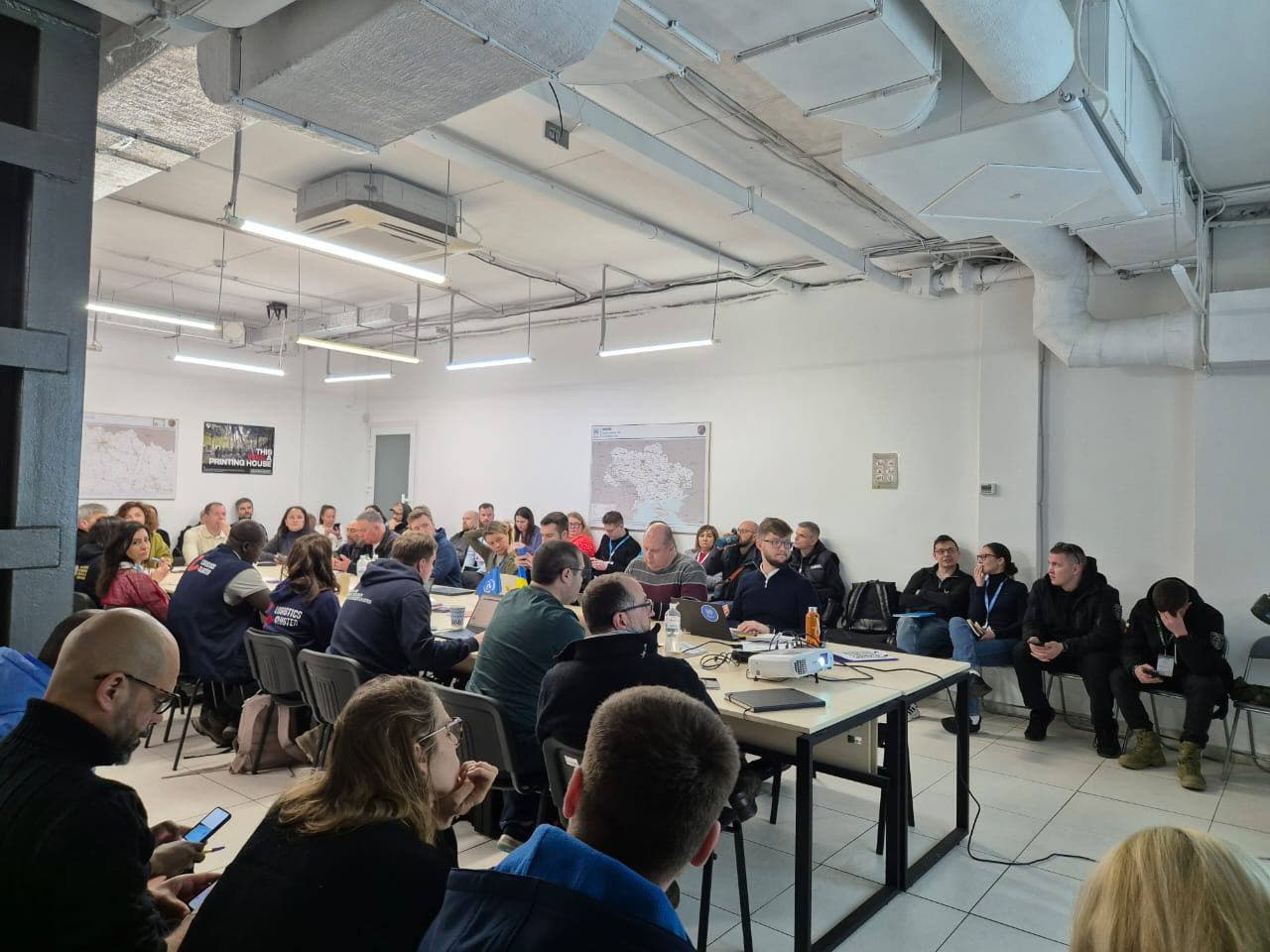 Велика група людей зібралася у конференц-залі з сучасною індустріальною стелею, що має відкриті труби та вентканали. Столи розташовані у формі літери "U", а учасники користуються ноутбуками, роблять нотатки чи переглядають документи. На стінах розміщені карти та постери, а на одному зі столів стоїть проектор, що вказує на презентацію чи семінар.