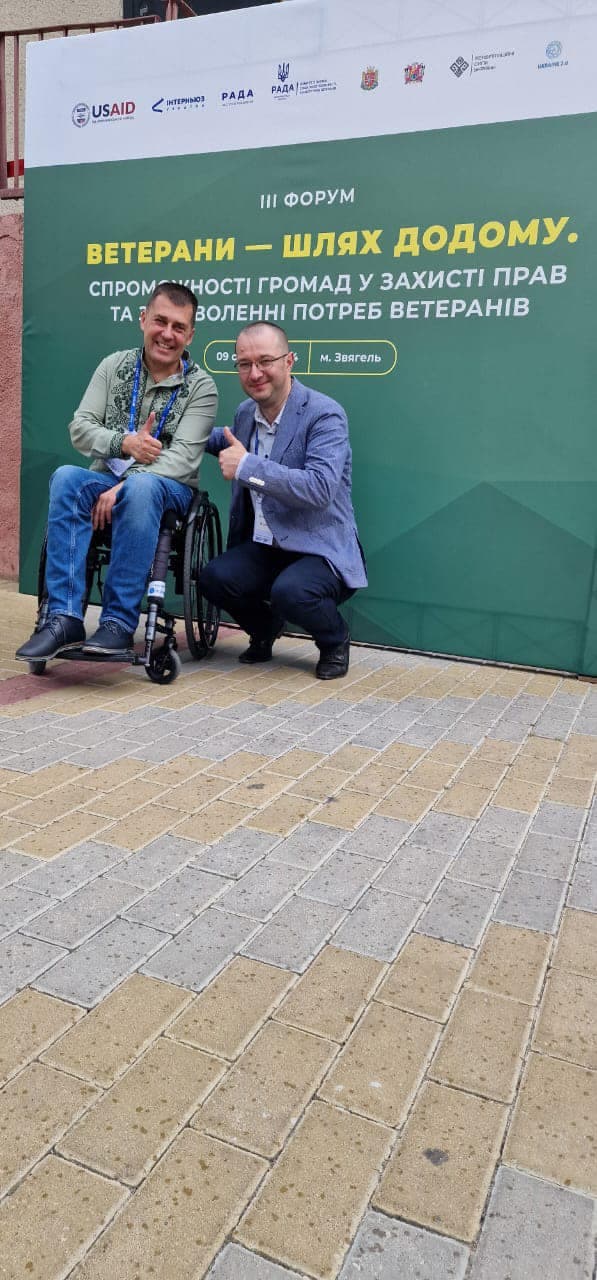 Два чоловіки сидять на фоні зеленого банера.