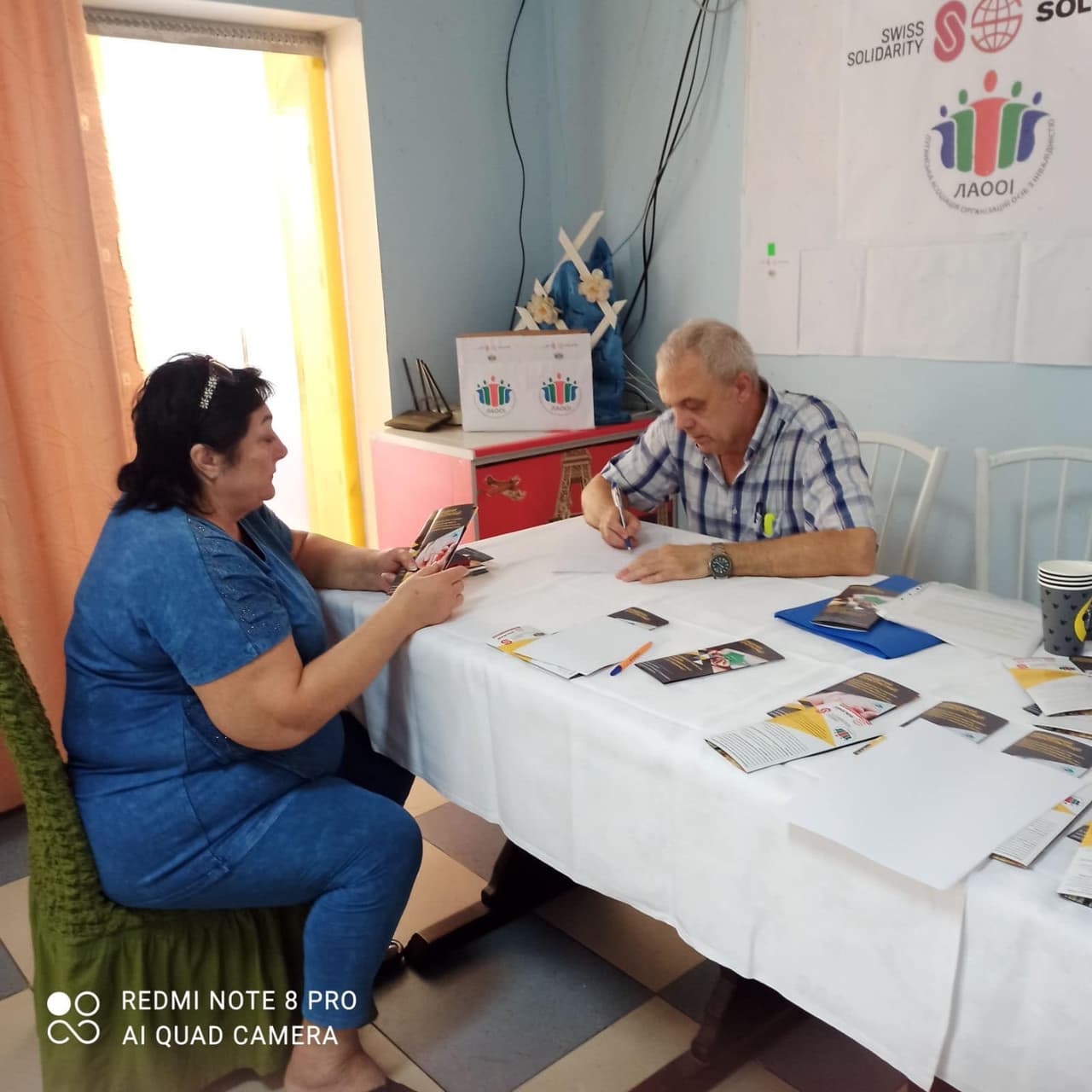 Чоловік сидить за столом і пише, напроти нього сидить жінка, яка тримає в руці флаєр.