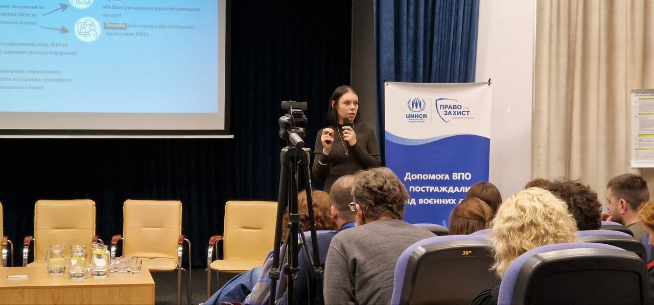 Presentation on Assistance for IDPs: A woman in black clothing speaks to an audience during a conference, holding a microphone. In the background, a screen displays a presentation, and a banner with the logos of UNHCR and “Right to Protection” contains the text “Assistance for IDPs, affected by military actions.” The audience is present in the hall, with some attendees taking notes.