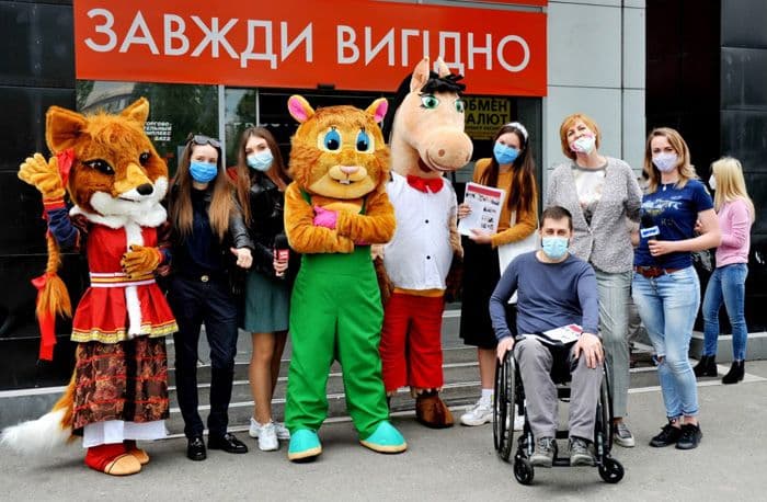 A group of people wearing protective masks pose for a photo in front of a building with the sign "ALWAYS PROFITABLE." Among them are two individuals in fairy-tale character costumes – a fox and a cat, a person in a wheelchair, and women holding leaflets. The atmosphere is friendly and positive.