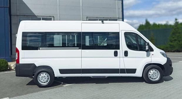 A white passenger van with a long body and multiple side windows is parked on a paved surface near a building with a metallic exterior. The van features a sliding side door and standard wheels. In the background, there is a building with large windows and a green area with trees. The scene shows a vehicle designed to carry several passengers.