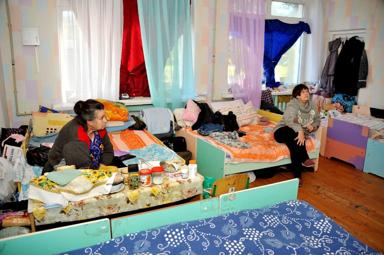 Two women are in a room arranged as a temporary shelter. Beds with personal belongings, a table with food, and colorful curtains on the windows.