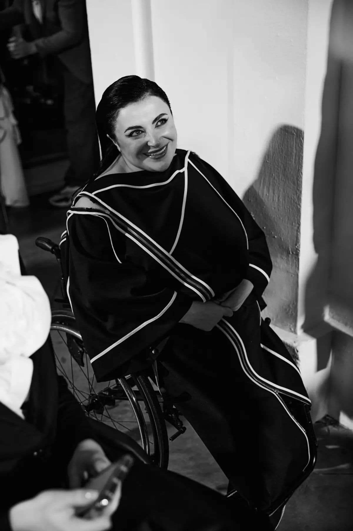 Black-and-white photograph: a person in a wheelchair sits near a wall, smiling with hands clasped in their lap. They wear a dark garment with white trim resembling a ceremonial or academic robe. In the foreground, another individual is partially visible holding a phone. The scene appears to capture a formal event or ceremony, emphasizing inclusion and representation of people with disabilities in celebratory contexts.