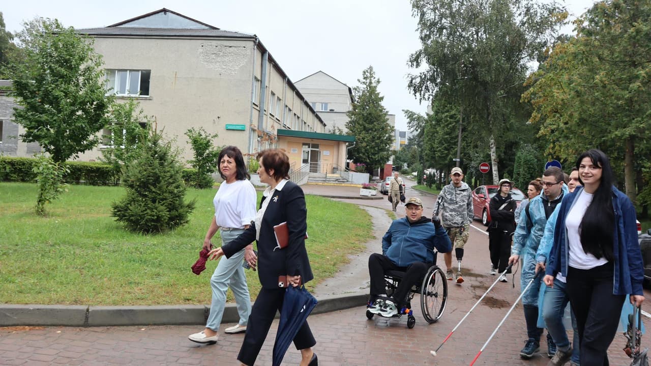 Група людей йде по вимощеній доріжці на відкритій території з будівлями на фоні. Серед них — особи з білими тростинами та одна людина у візку, яких супроводжують асистенти. Навколо — дерева, трава та багатоповерхова будівля з ознаками зношеності, що нагадує територію кампусу або парку в житловому районі. Сцена підкреслює доступність і інклюзивність для людей з порушеннями зору та мобільності у громадському просторі.